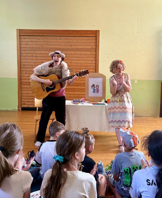 Eine Gruppe Schüler sitz vor der vorlesenden Autorin und einem Gitarrenspieler